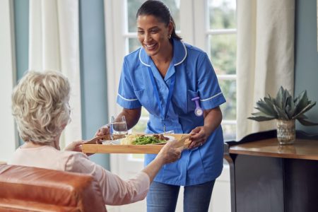 female-care-worker-in-uniform-bringing-meal-on-tray-to-senior-woman-sitting-in-lounge-at-home.jpg